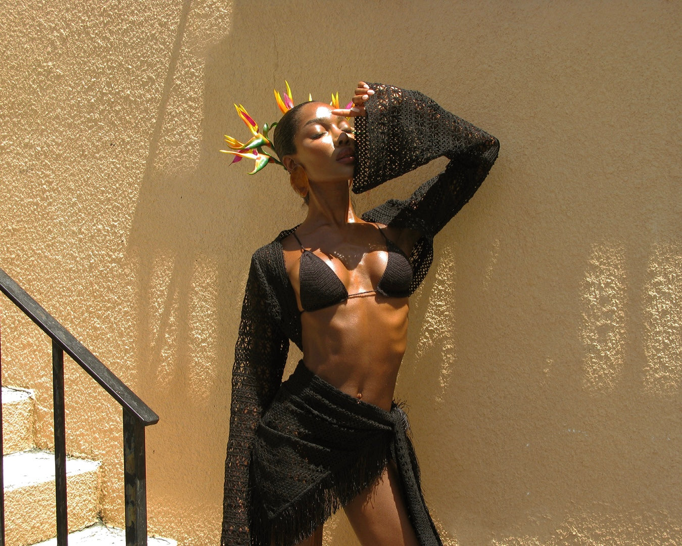 Captured in mid-pose, the model wears the Sol Bikini and Chica Bolero with the Palama Sarong wrapped tightly at the hips. Her arms frame her face as she looks upward into the sun, the bright floral headpiece adding vibrance to the warm-toned setting.