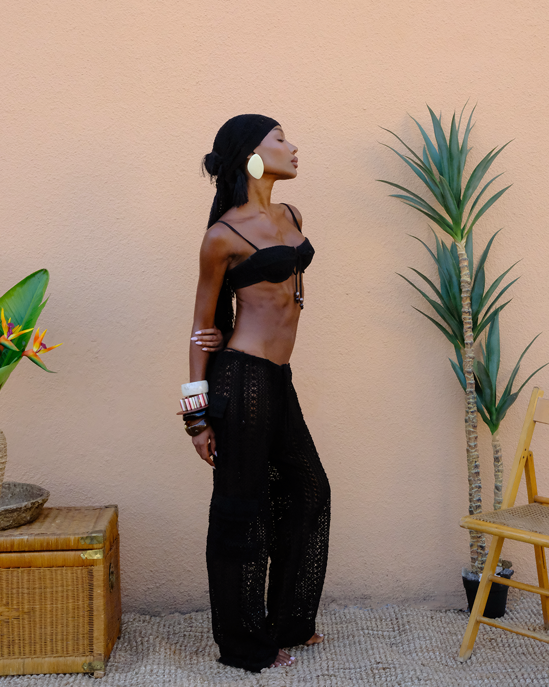 Model wearing Heart of Sun’s black crochet look featuring the Dolce Vita Bikini top, Bahia Cargo pants, and Palama Sarong. Styled in a minimal tropical setting with woven textures, rattan accents, and bold accessories for a luxurious resortwear vibe.