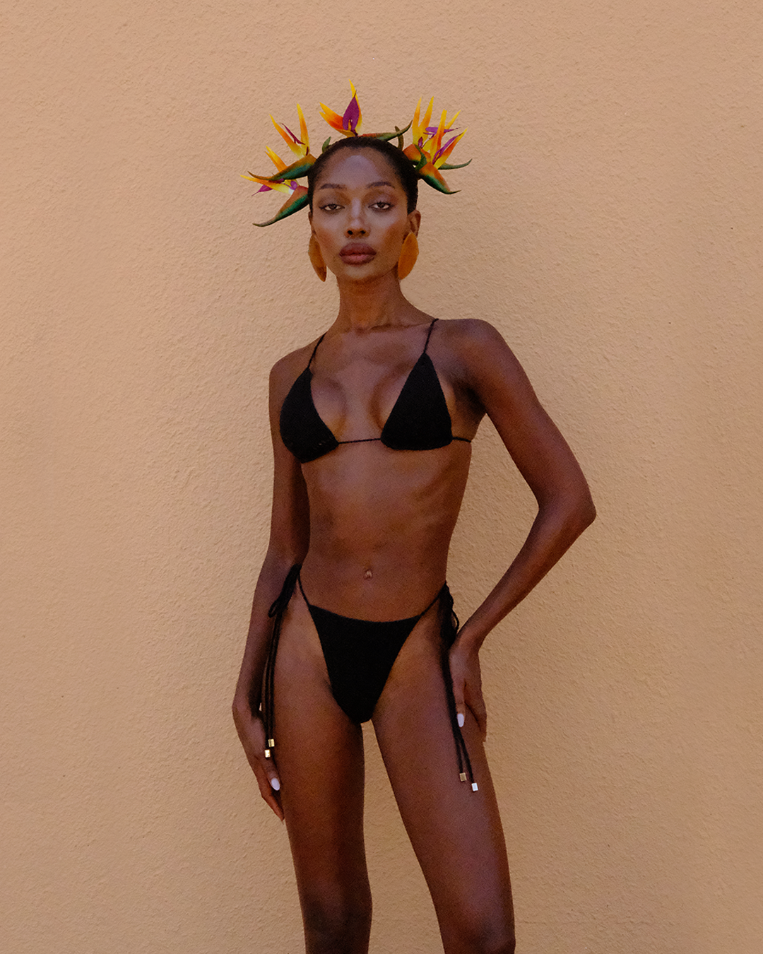 Model poses confidently in the Sol Bikini, featuring a sleek black triangle top and string bottoms with gold hardware. A bold tropical floral headpiece and large wooden earrings accent the look against a sunlit beige wall.