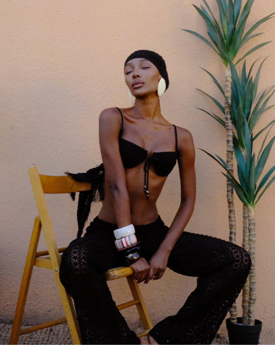 Heart of Sun’s signature crochet set — Dolce Vita Bikini, Palama Sarong, and Bahia Cargo — styled on model sitting on a wooden chair in a relaxed tropical setting with woven textures and palm accents.