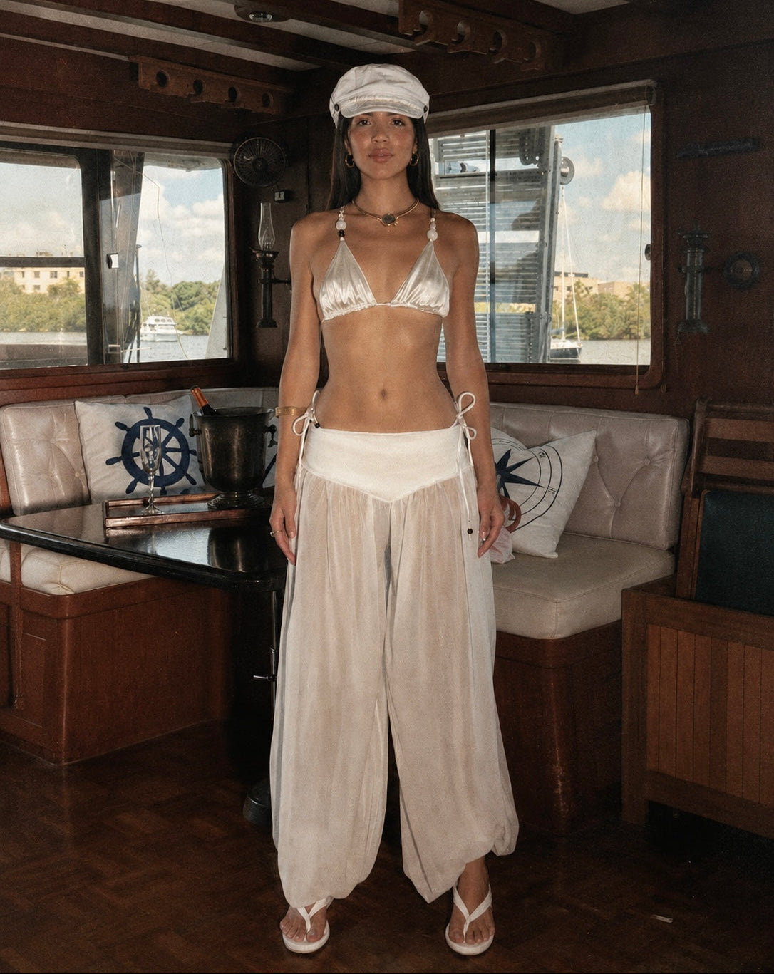 A woman stands inside a wooden boat cabin wearing a white nautical-themed two-piece swimsuit with a halter top, matching head covering, and loose wide-leg bottoms.